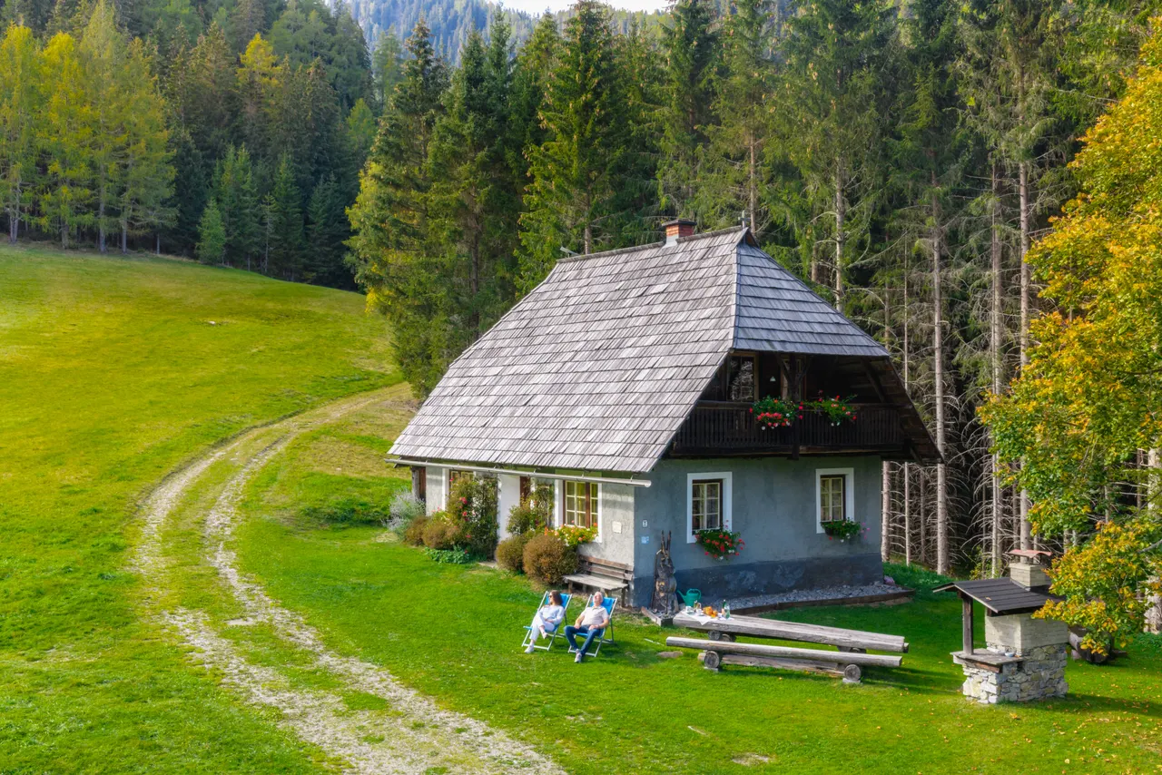 Die ganze Hütte, davor zwei Menschen in Liegestühlen, Holztisch und gemauerter Grill