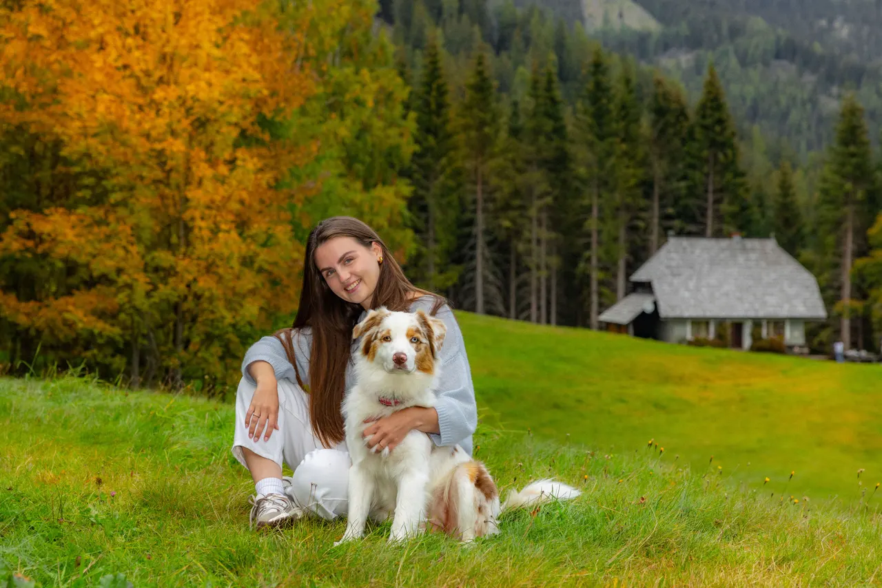 Eine junge Frau mit dem Hund auf der Wiese, dahinter die Hütte